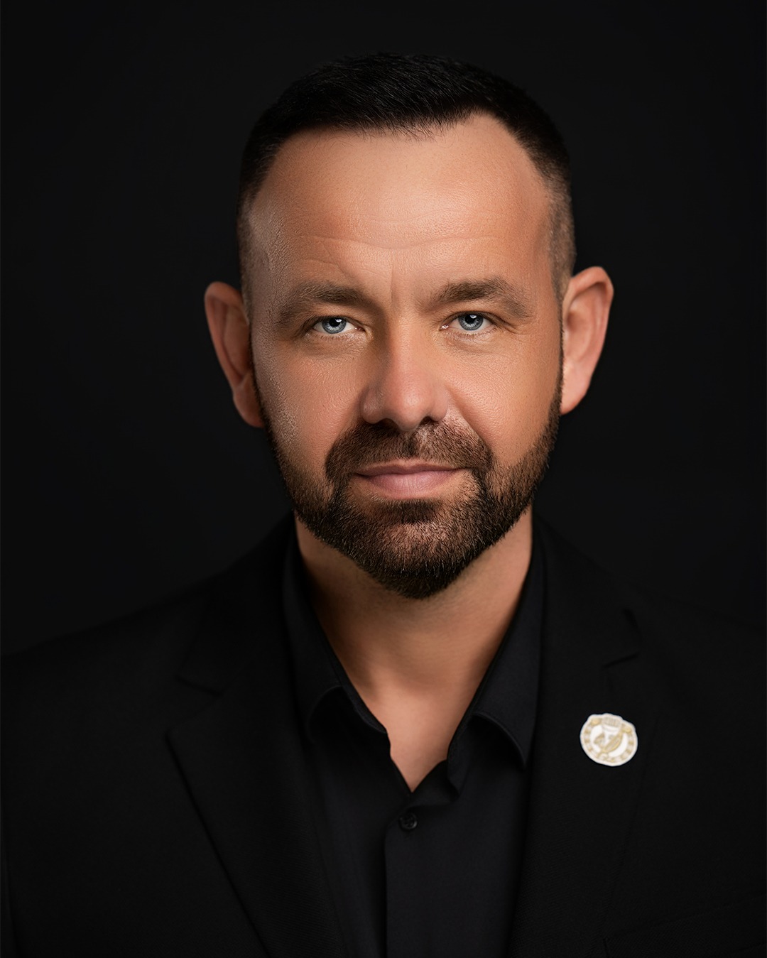 Mężczyzna w czarnym garniturze i koszuli, z brodą, patrzący wprost. Na klapie marynarki ozdobna przypinka. Portret na ciemnym tle, oświetlenie studyjne.