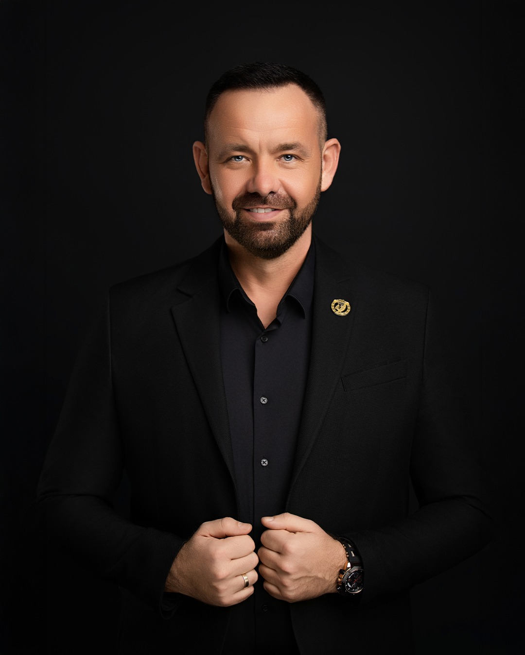 Portret biznesowy mężczyzny w czarnym garniturze na ciemnym tle, z delikatnym uśmiechem i widoczną odznaką w klapie marynarki. Zegarek na nadgarstku.