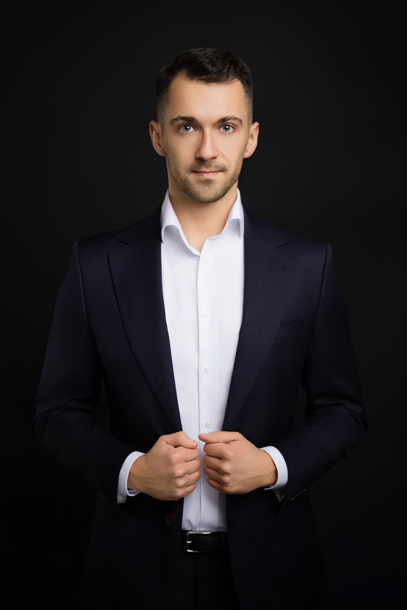 Portret mężczyzny w garniturze na czarnym tle, z delikatnym uśmiechem i jasnymi oczami, trzymającego dłonie na klapach marynarki, oświetlonego studyjnym światłem.