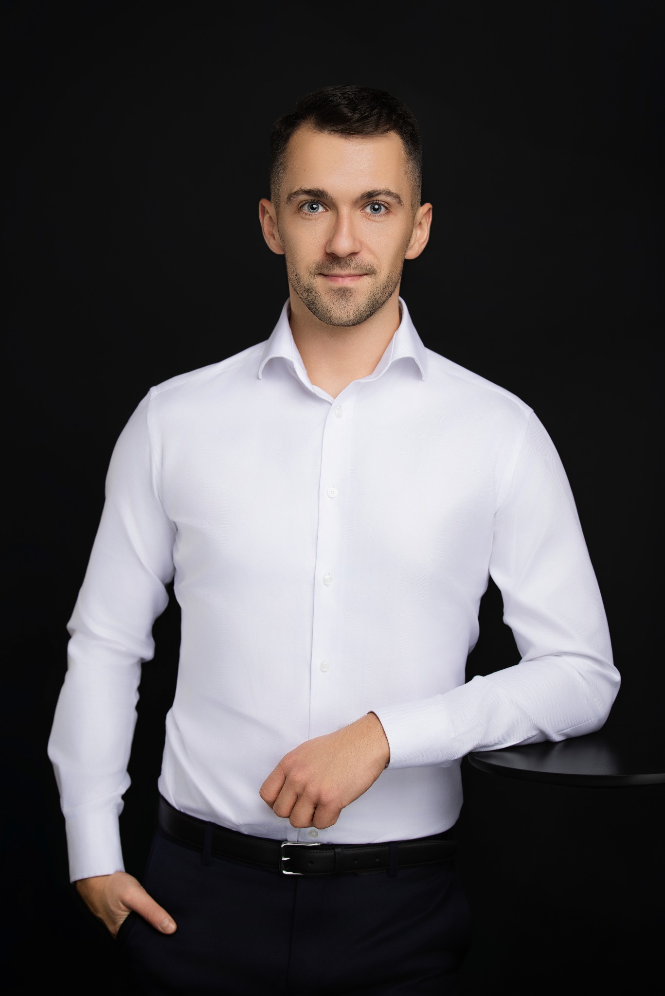 Mężczyzna w białej koszuli, oparty o czarny stolik na ciemnym tle. Profesjonalny portret biznesowy z subtelnym uśmiechem i wyraźnymi oczami.