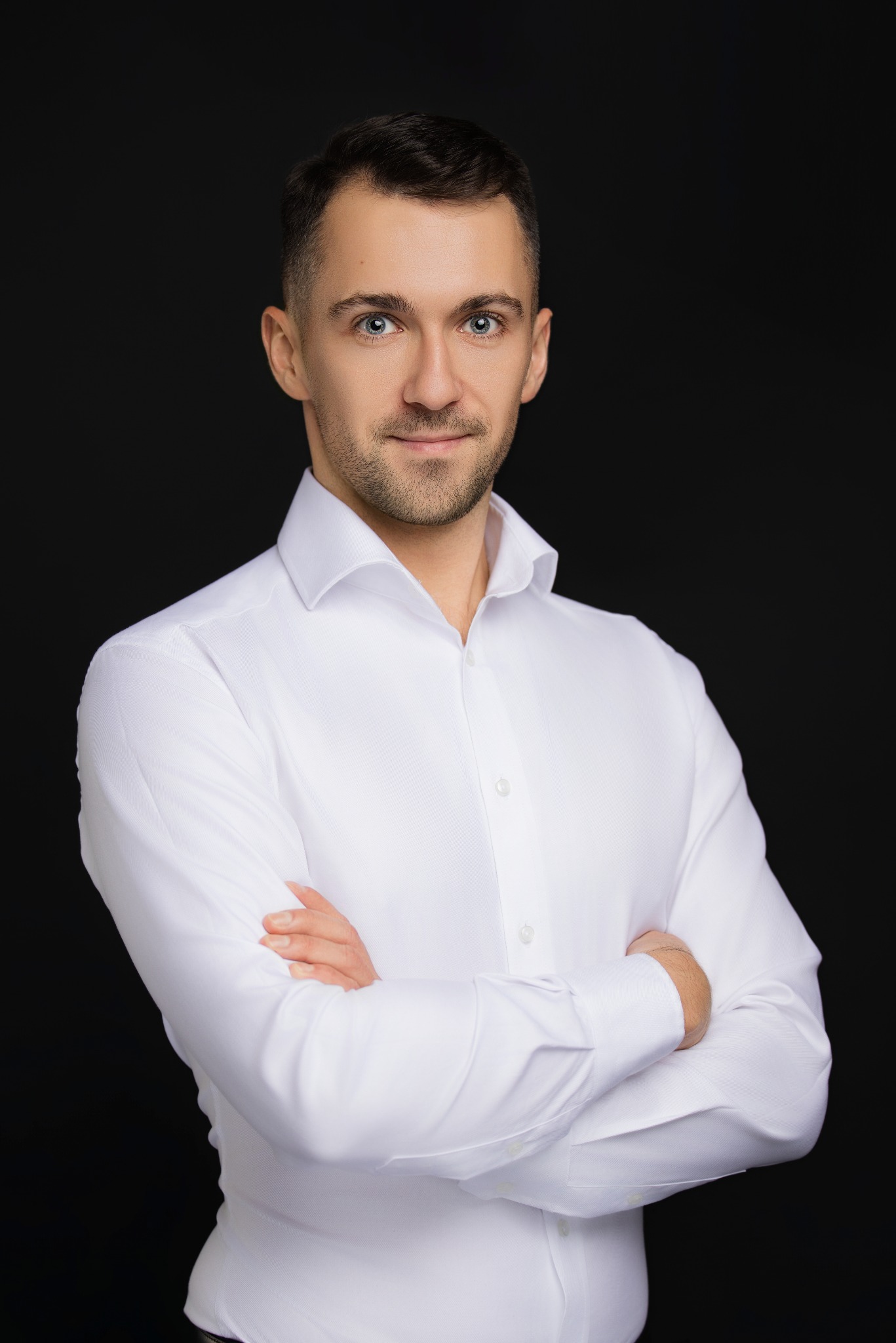 Portret biznesowy mężczyzny w białej koszuli na czarnym tle, z założonymi rękami i lekkim uśmiechem, ukazujący profesjonalizm i pewność siebie.