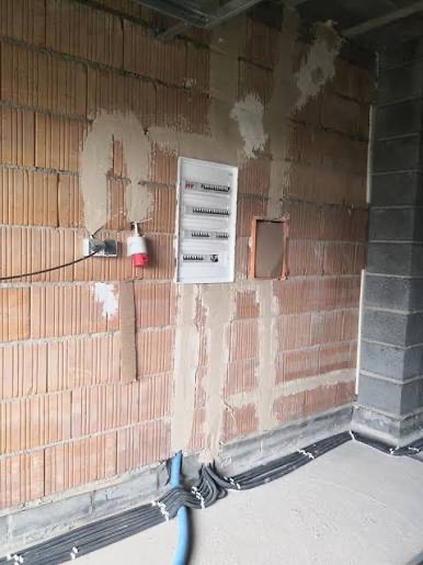 Rozdzielnica elektryczna zamontowana na ceglanej ścianie w surowym stanie, widoczne kable w peszlach przy podłodze i gniazdo siłowe. Prace instalacyjne w trakcie.