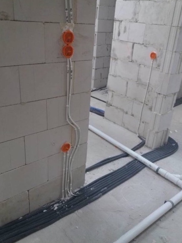 Instalacja elektryczna w surowym budynku: pomarańczowe puszki podtynkowe i białe przewody na ścianie z bloczków, czarne rury karbowane na podłodze.