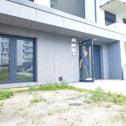 MPV Group Sp. z o.o. - Nowoczesna elewacja budynku z szarymi panelami i dużymi oknami. Otwarta szklana witryna z widoczną osobą w środku. Minimalistyczny styl, jasne otoczenie.