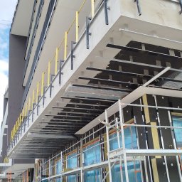 MPV Group Sp. z o.o. - Budynek w trakcie ocieplania: rusztowania, izolacja z wełny mineralnej, folia ochronna na oknach. Widok z dołu na elewację w trakcie prac.