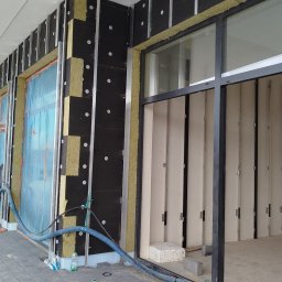 MPV Group Sp. z o.o. - Ocieplanie budynku z widoczną warstwą izolacyjną i elementami montażowymi. Fragment elewacji z oknem w trakcie prac wykończeniowych.