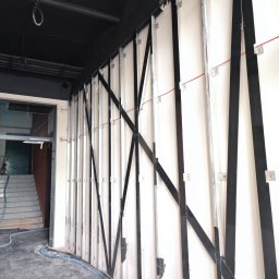MPV Group Sp. z o.o. - Wnętrze w trakcie prac: białe płyty mocowane do metalowej konstrukcji na ścianie, czarny sufit z instalacjami. Widoczne elementy przygotowania do dalszych prac wykończeniowych.