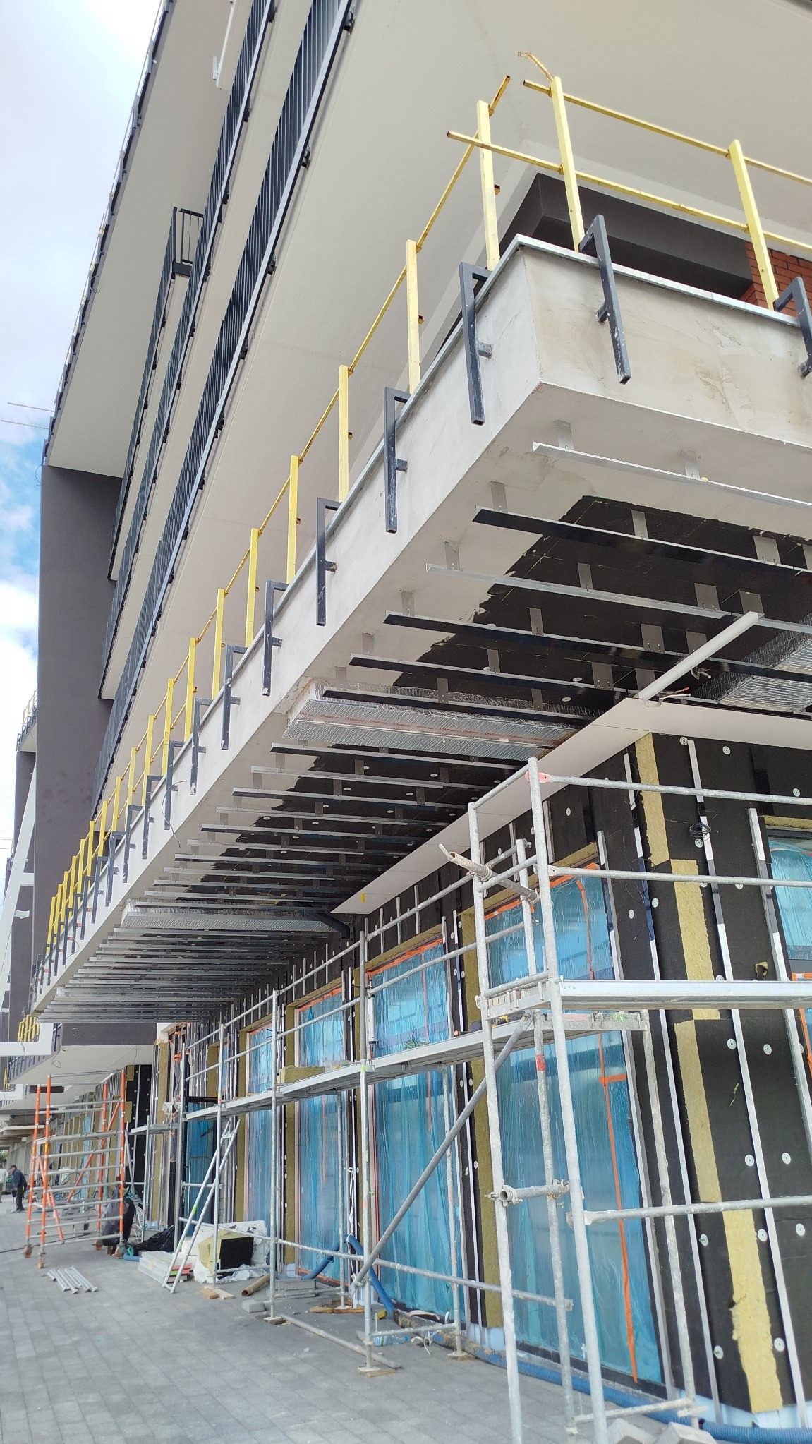 Budynek w trakcie ocieplania: rusztowania, izolacja z wełny mineralnej, folia ochronna na oknach. Widok z dołu na elewację w trakcie prac.