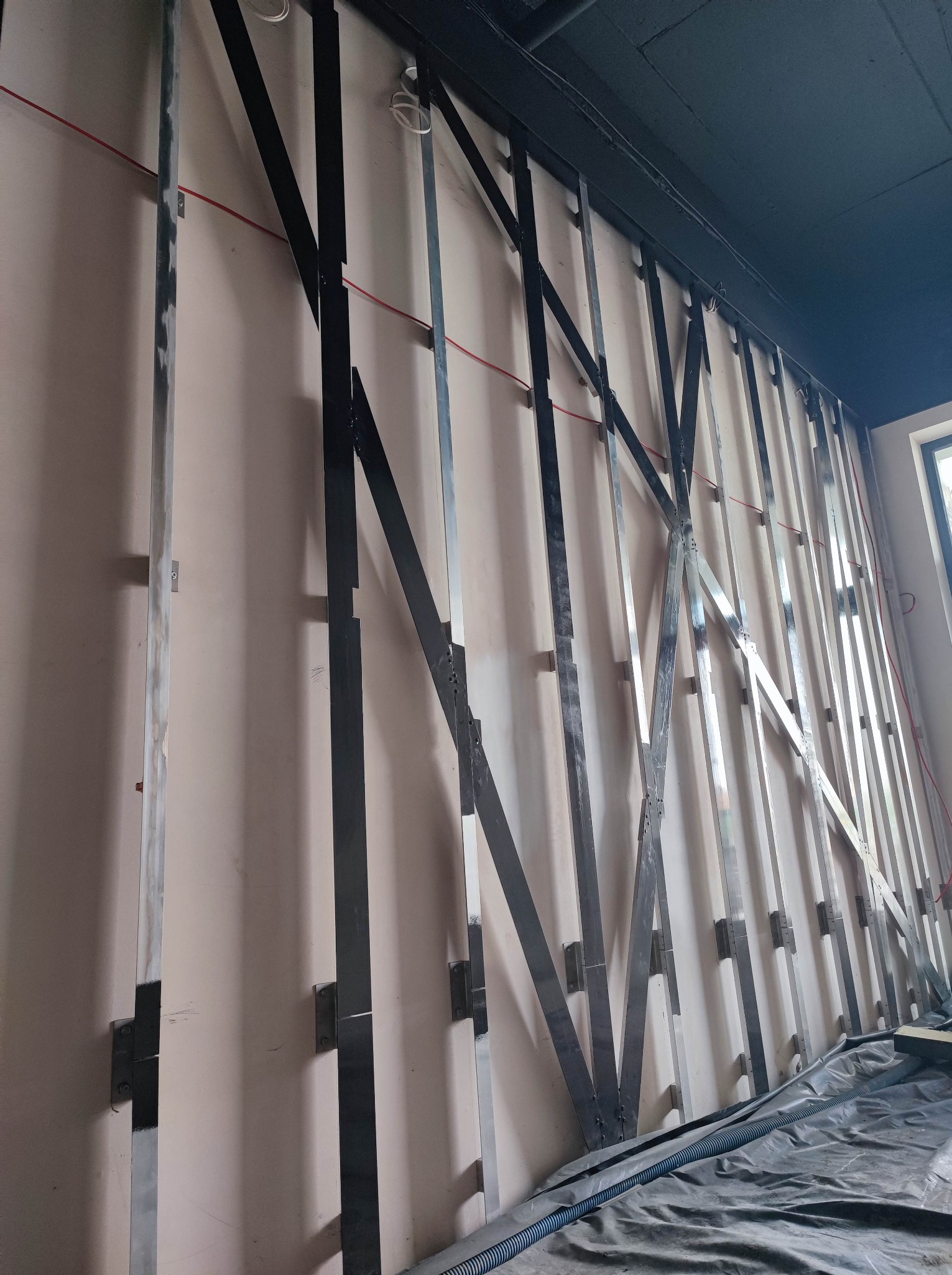 Ściana z zamontowaną metalową konstrukcją szkieletową z czarnymi i srebrnymi profilami, przygotowana pod ocieplenie. Widoczne elementy instalacji.