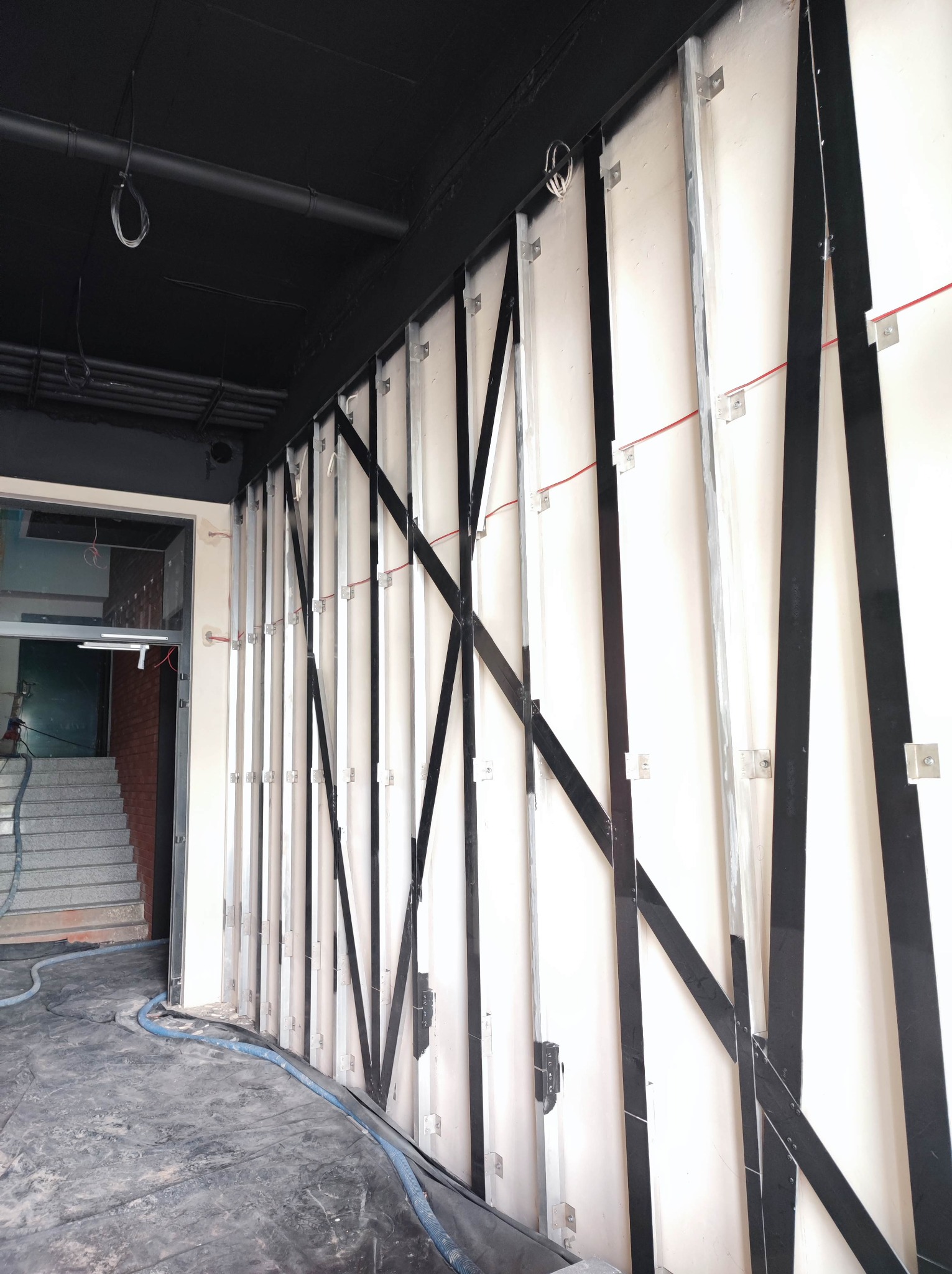 Wnętrze w trakcie prac: białe płyty mocowane do metalowej konstrukcji na ścianie, czarny sufit z instalacjami. Widoczne elementy przygotowania do dalszych prac wykończeniowych.