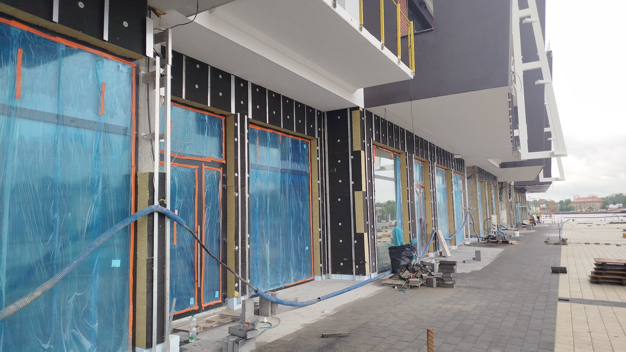 Montaż izolacji termicznej na ścianie budynku z oknami zabezpieczonymi folią. Widoczne rusztowania i materiały budowlane, sugerujące etap prac wykończeniowych.