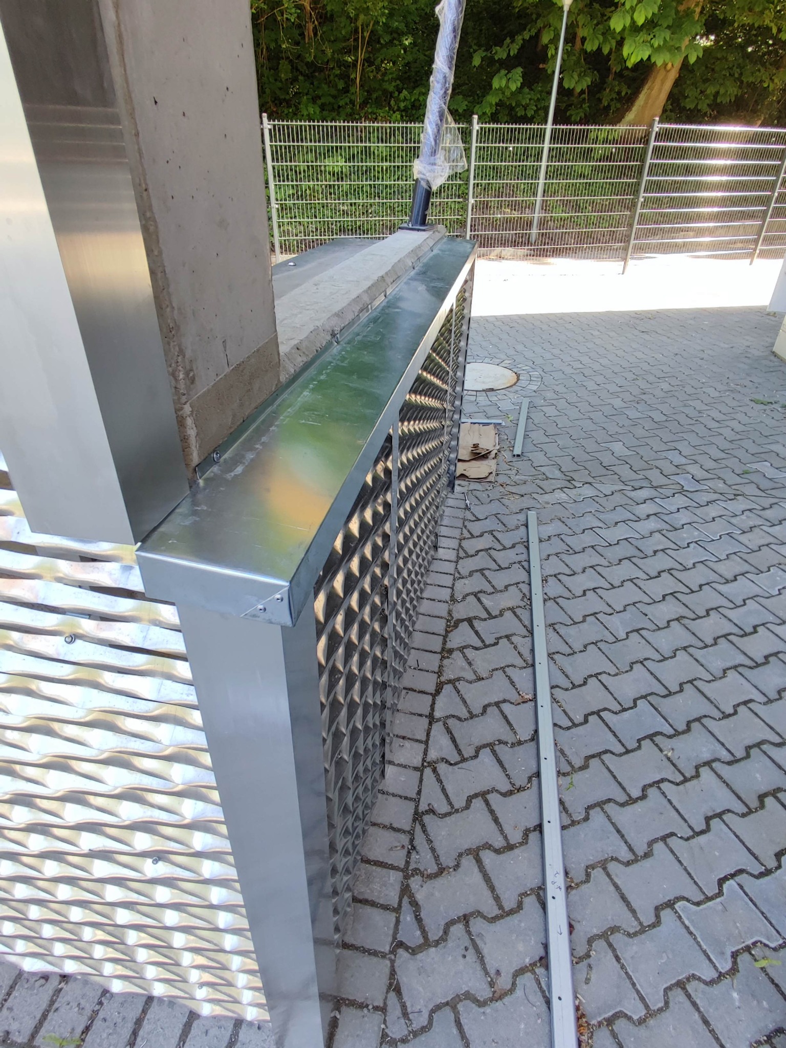 Montaż metalowej elewacji z perforowanymi panelami i obróbkami blacharskimi. Widoczne narzędzia i fragment chodnika z kostki. Częściowy widok ogrodzenia.