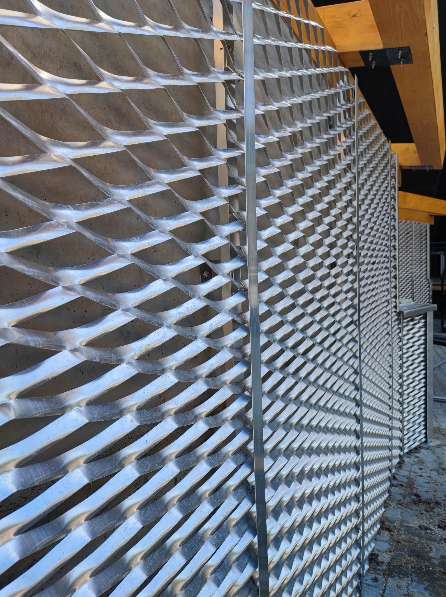 Metalowa elewacja budynku z ażurową kratą o romboidalnym wzorze, widok z dołu, fragment drewnianej konstrukcji dachu w tle.