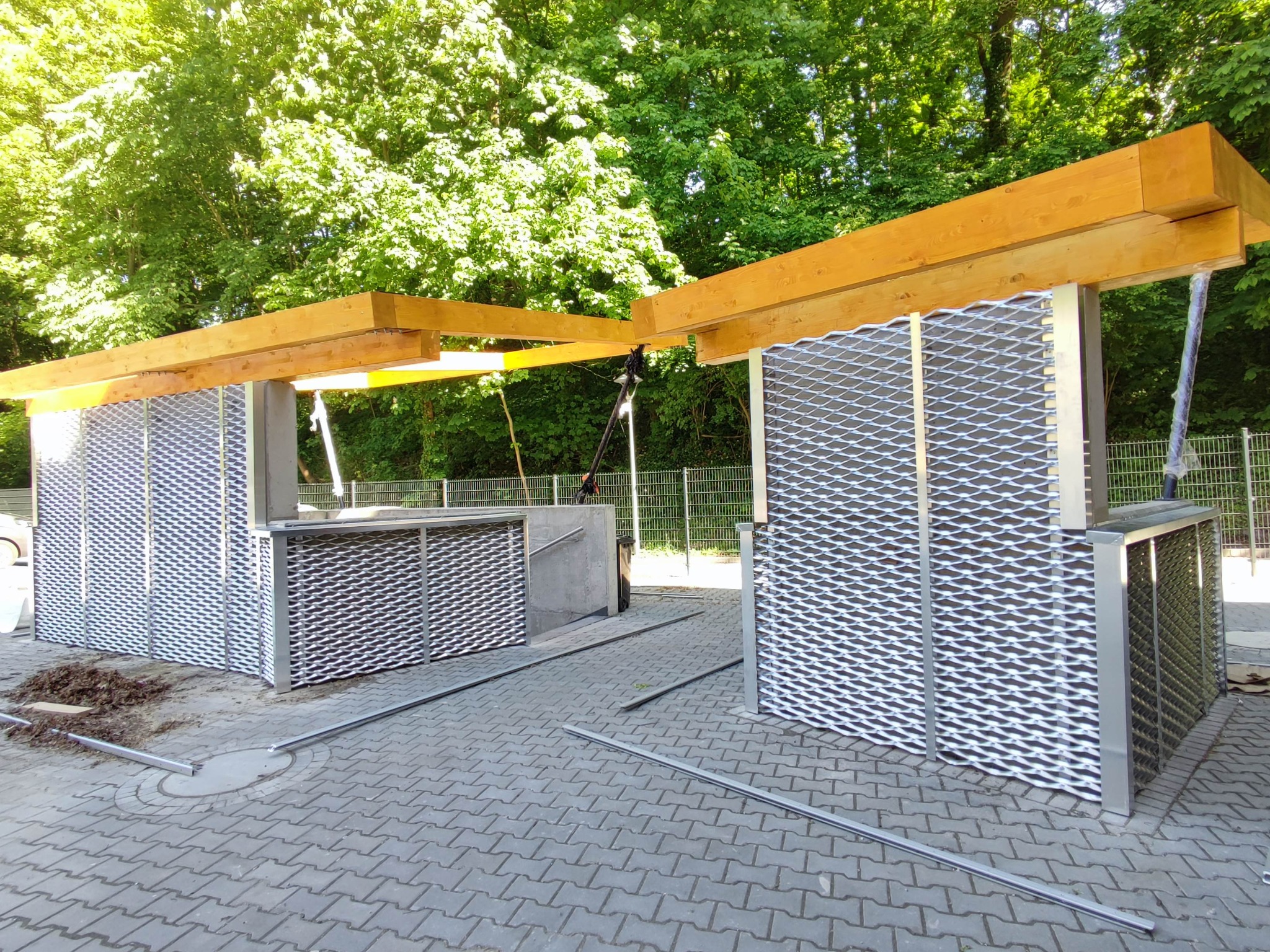 Nowoczesna elewacja z metalowej siatki i drewnianych belek, widok z zewnątrz na tle zieleni. Elementy konstrukcyjne na kostce brukowej.