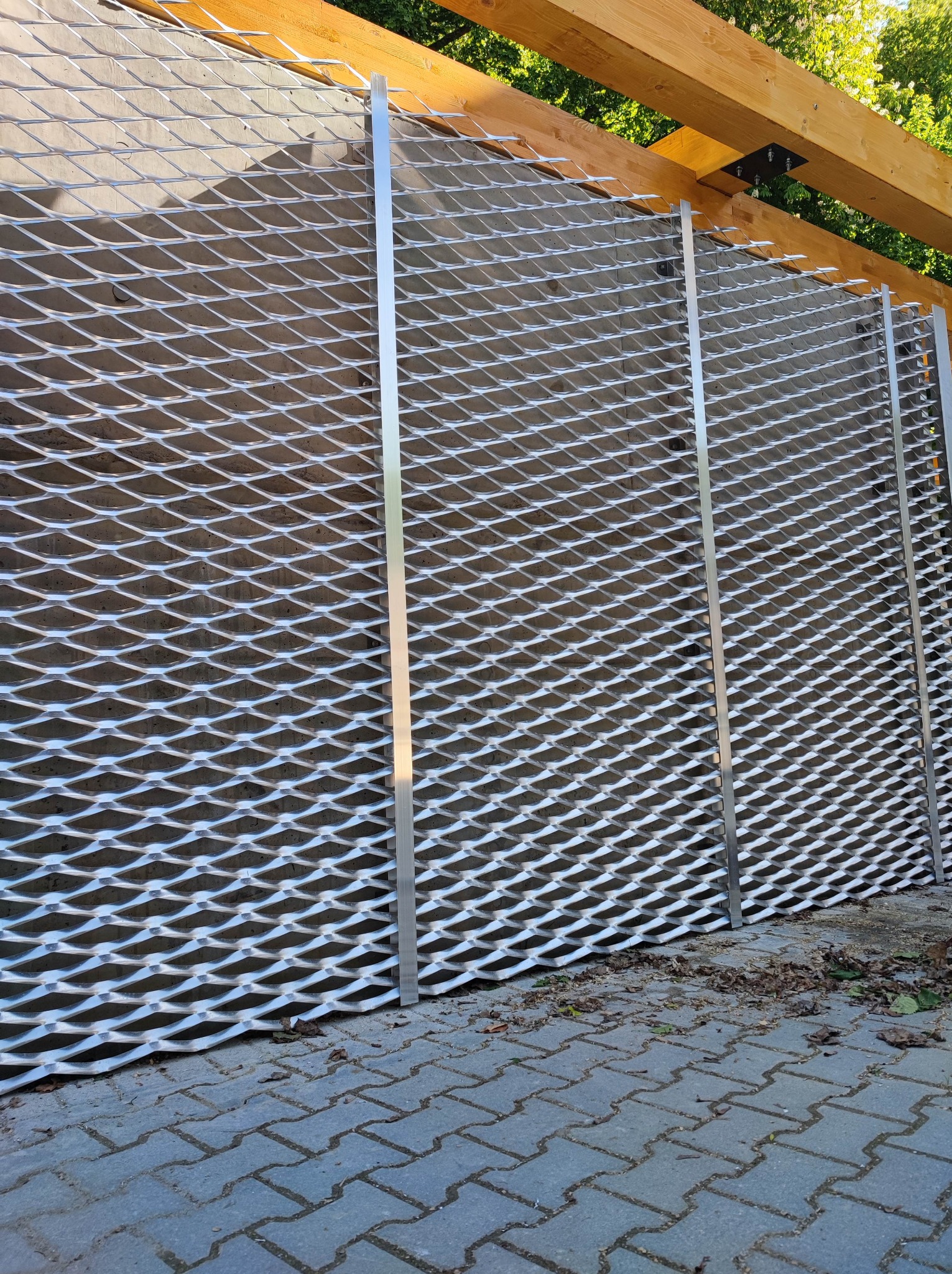 Montaż siatki elewacyjnej na betonowej ścianie z drewnianą konstrukcją wsporczą. Widoczne metalowe profile i regularny wzór siatki. Częściowo ułożona kostka brukowa.