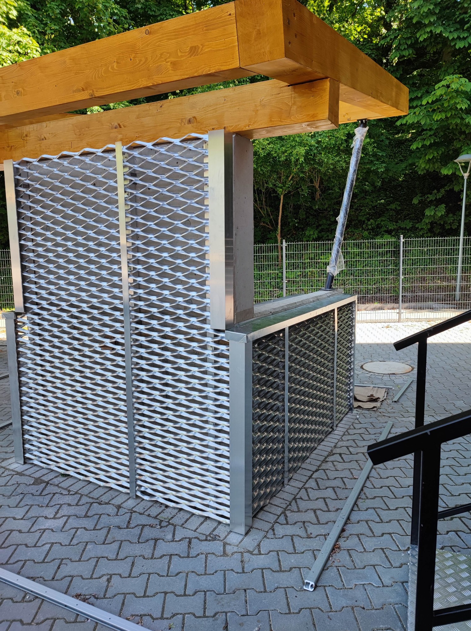 Konstrukcja z metalowej siatki i drewnianych belek, prawdopodobnie w trakcie montażu, na tle kostki brukowej i zieleni.