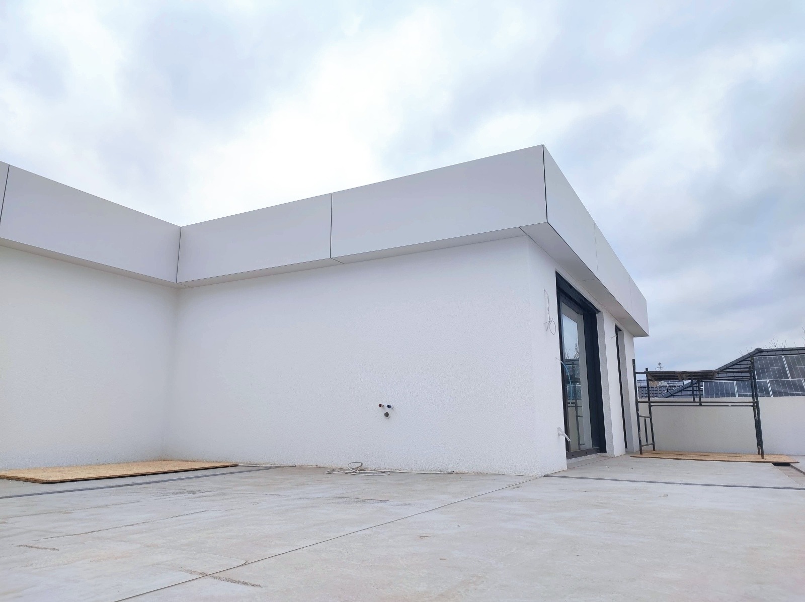 Biała elewacja budynku z panelami, widok z dołu, z fragmentem dachu sąsiedniego budynku z panelami słonecznymi w tle. Minimalistyczny design.