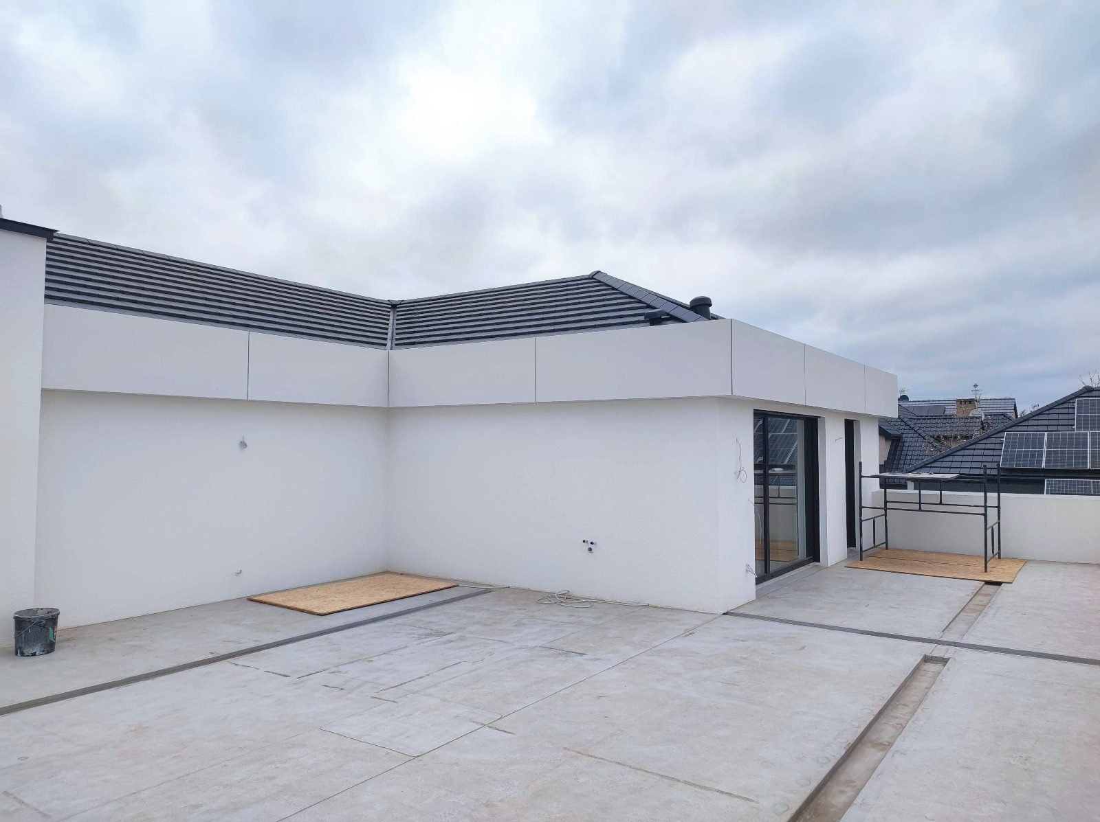 Biała elewacja budynku z widocznymi panelami, czarnym dachem i elementami wykończeniowymi. Częściowo wykończony taras z betonową posadzką. Widok na sąsiednie dachy z panelami słonecznymi.