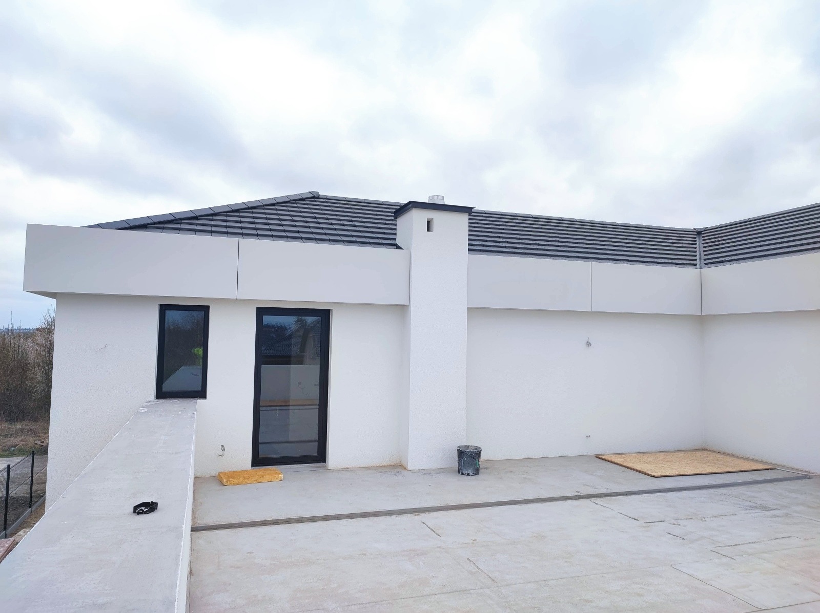 Nowoczesna elewacja domu z białym tynkiem, czarnymi ramami okiennymi i kominem. Widoczny fragment dachu z szarą dachówką. Ujęcie z poziomu tarasu.