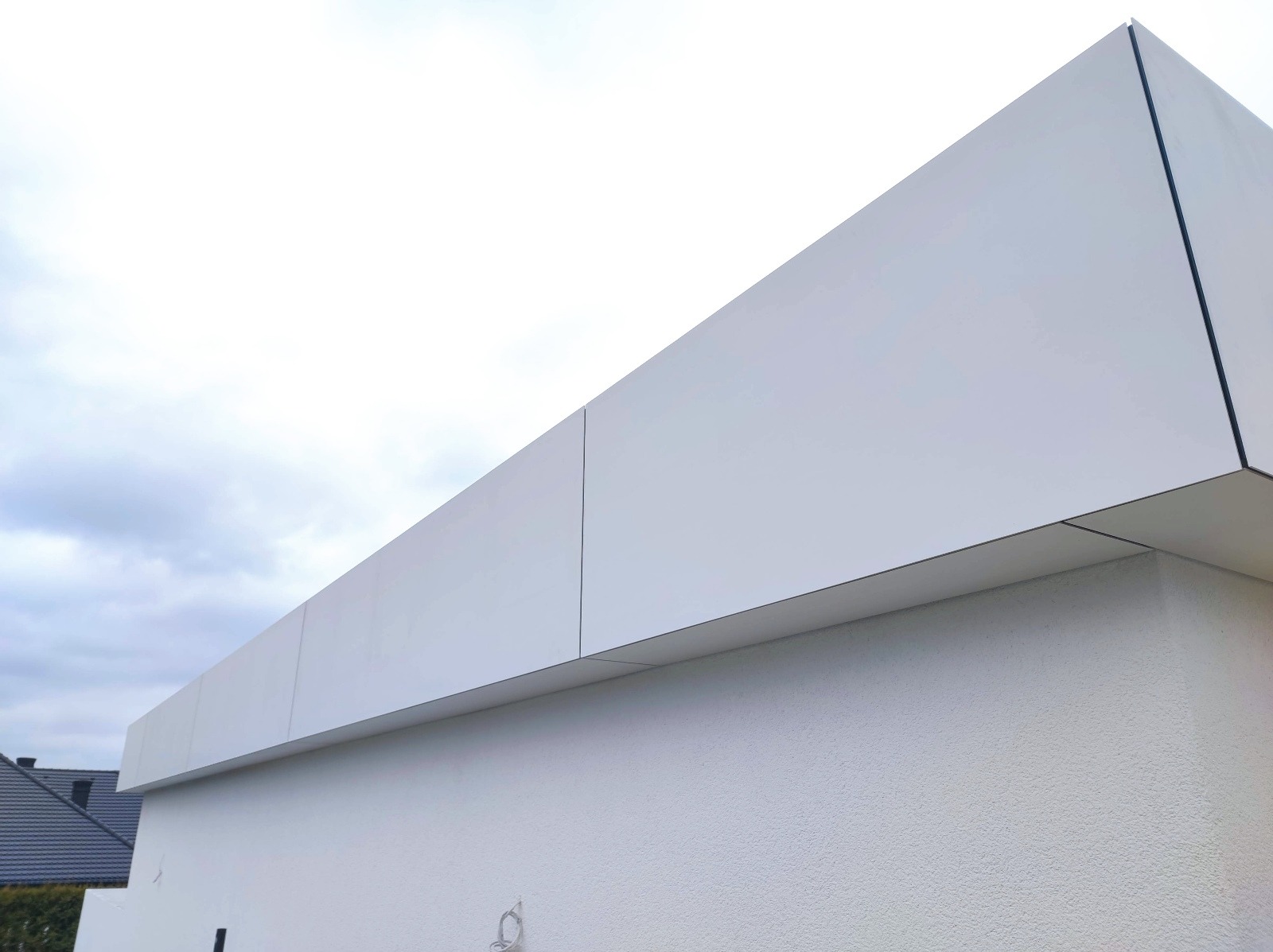 Nowoczesna, biała elewacja domu jednorodzinnego z widocznymi panelami elewacyjnymi i fragmentem dachu sąsiedniego budynku na tle pochmurnego nieba.