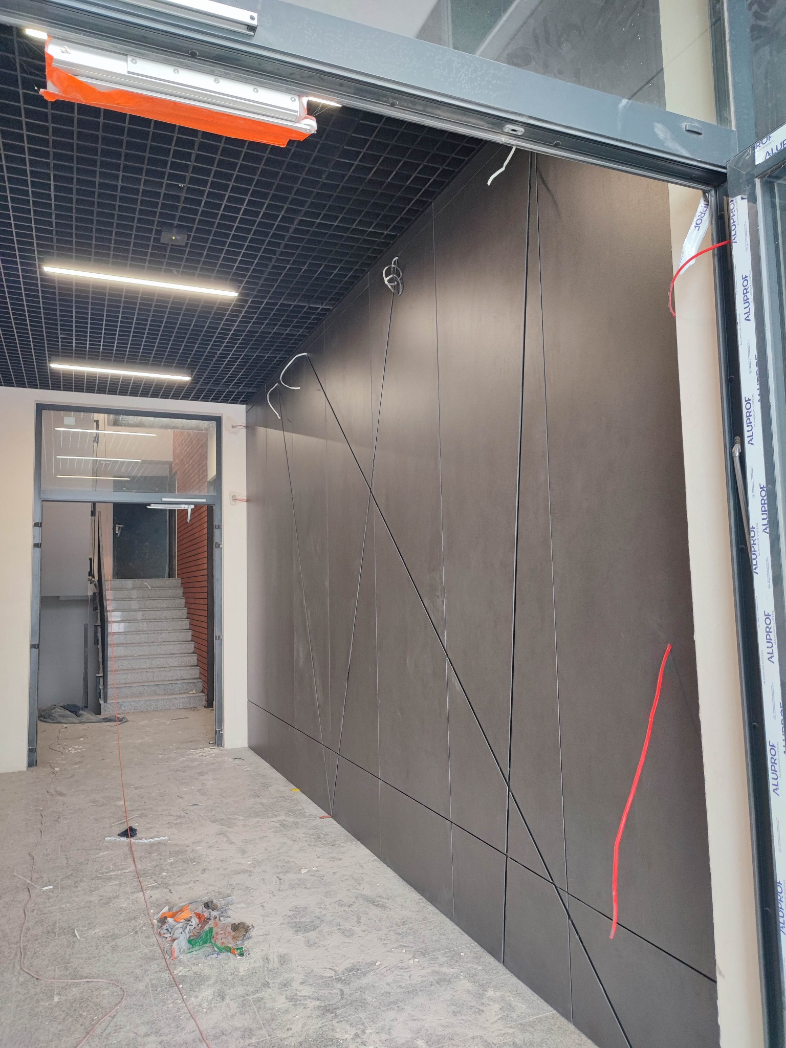 Nowoczesna elewacja wewnętrzna z paneli w odcieniach szarości, ułożonych w geometryczny wzór. Widoczne fragmenty instalacji elektrycznej i wejście na schody.