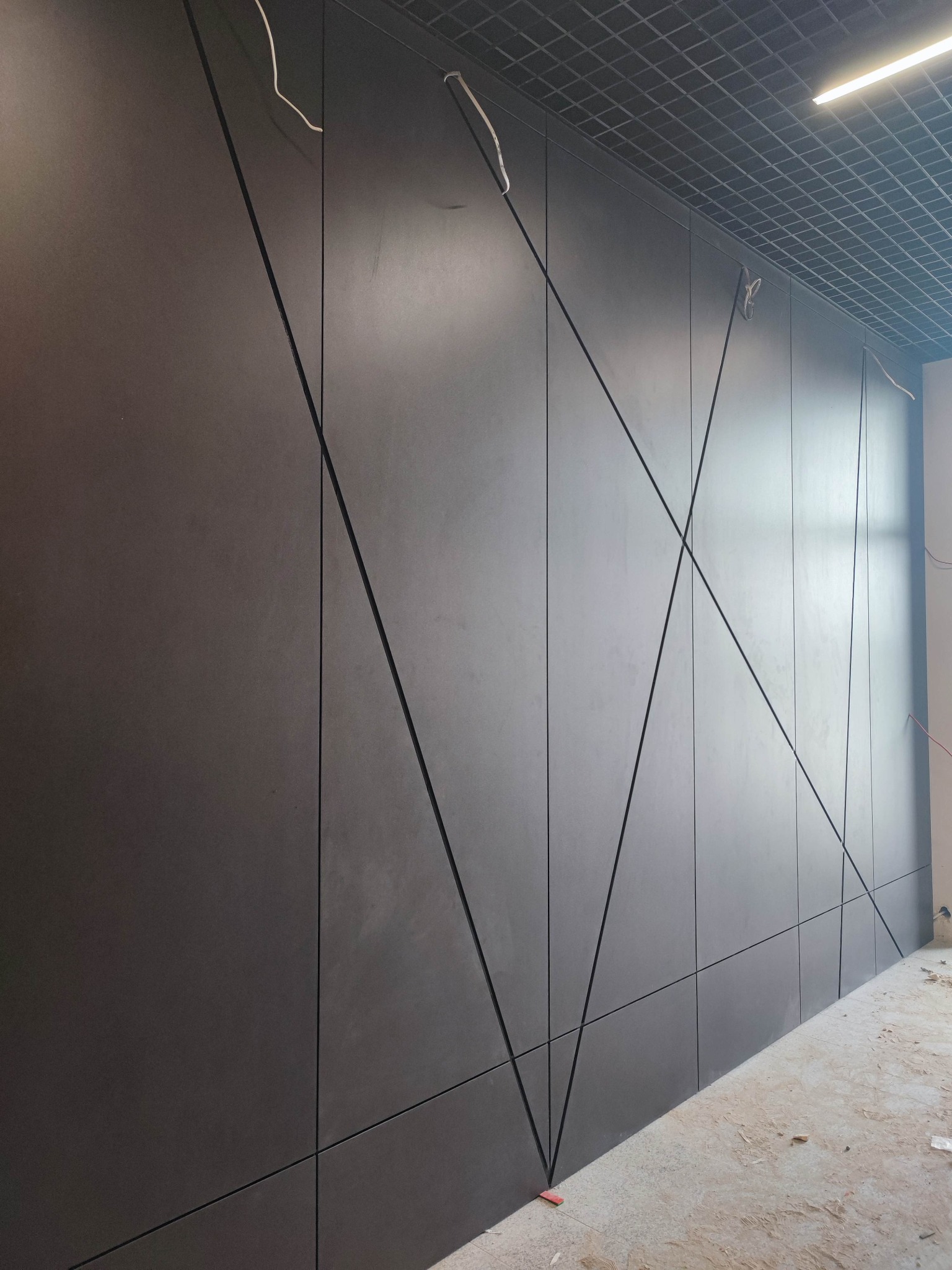 Grafitowa elewacja wewnętrzna z geometrycznym wzorem, podkreślonym czarnymi listwami. Widoczne przewody i fragment sufitu rastrowego.