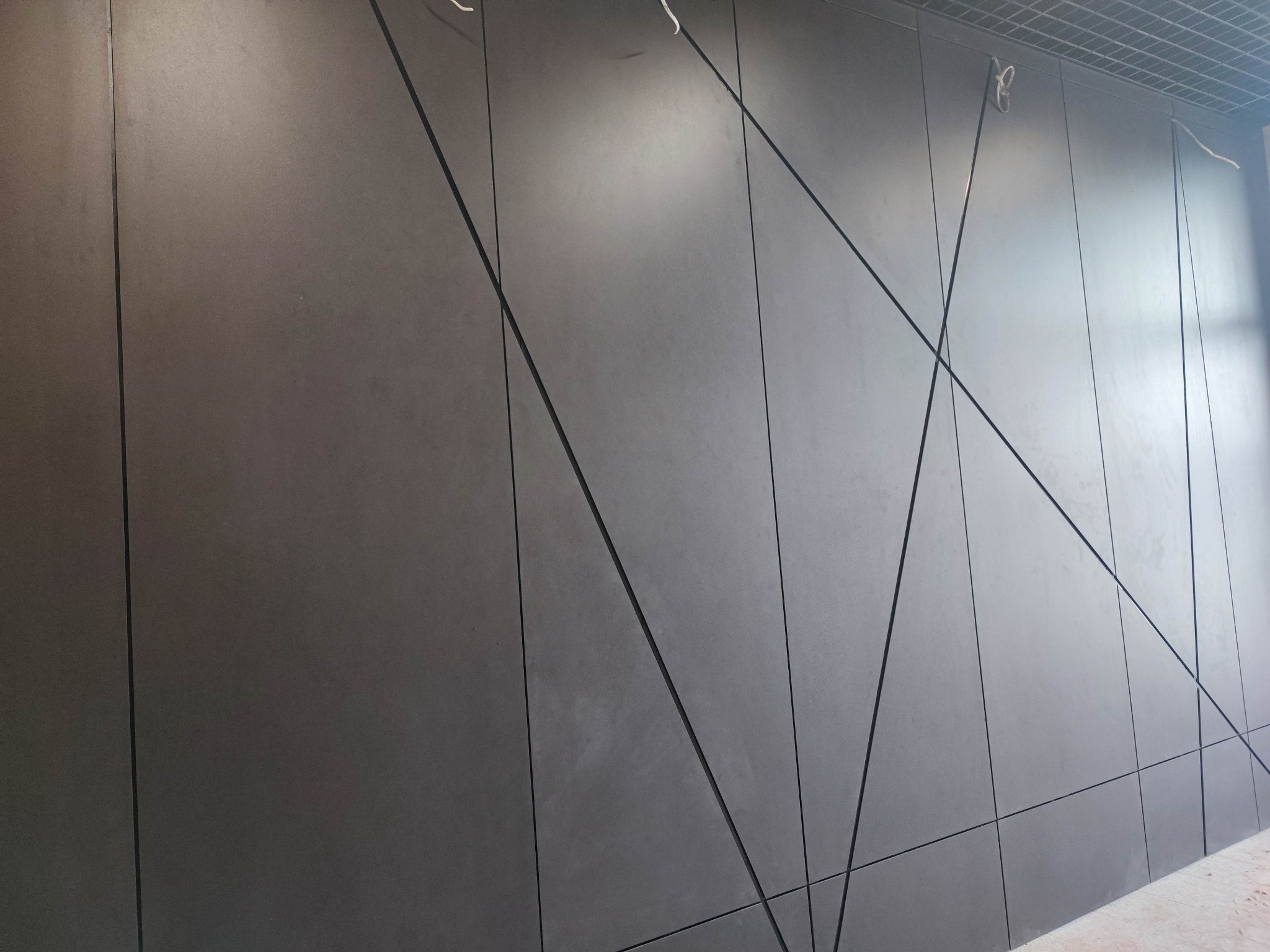 Grafitowa elewacja z paneli kompozytowych, ułożonych w geometryczny wzór. Widoczne łączenia paneli i przewody pod sufitem.