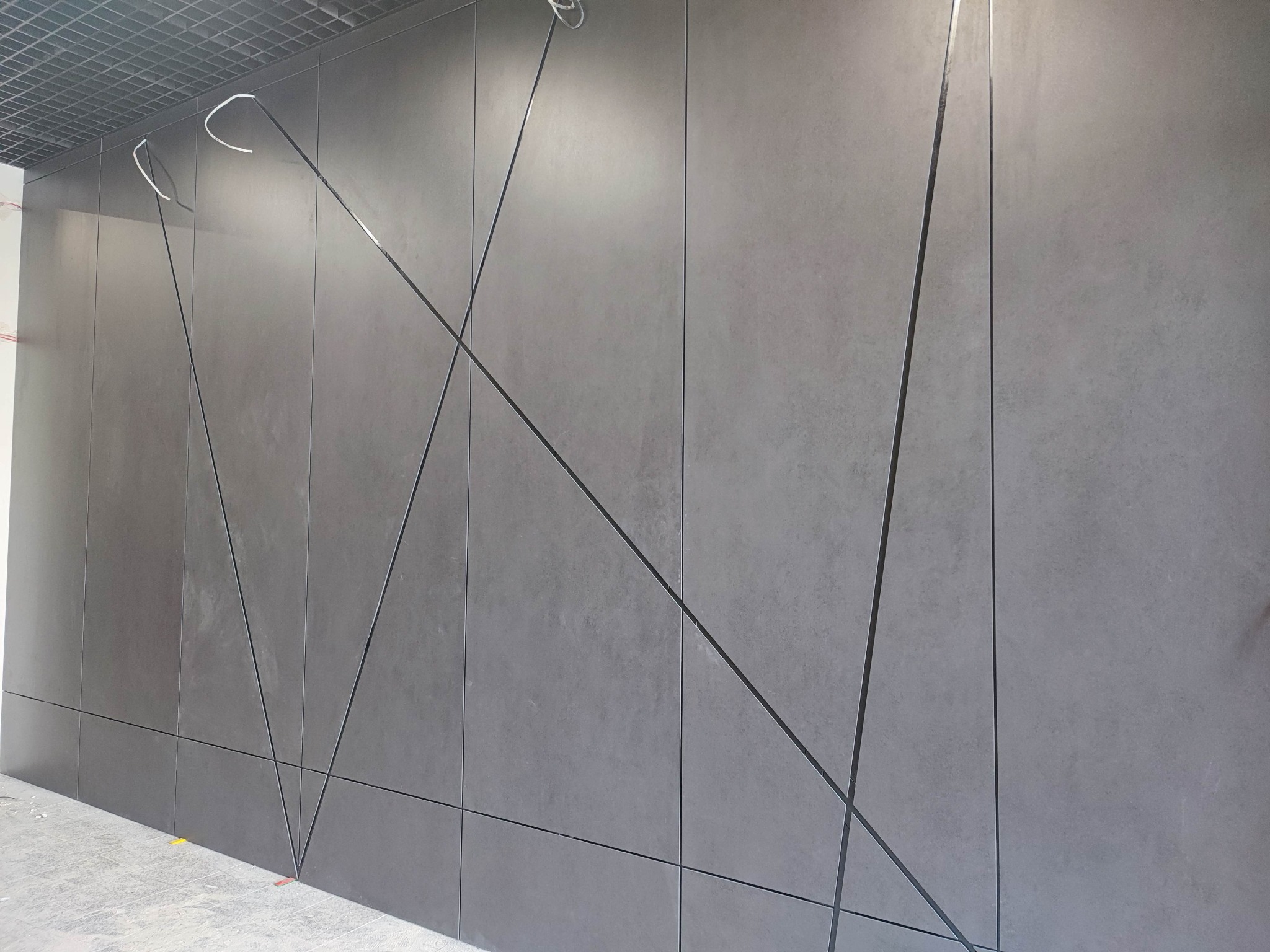 Szara elewacja z płyt z geometrycznym wzorem, widoczne kable pod sufitem, surowy stan wykończenia wnętrza.