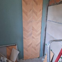 „Usługi Ogólnobudowlane KRIBUD” - Układanie płytek w jodełkę w Choczewie. Ściana w trakcie wykańczania, widoczne klej i płytki w ciepłych odcieniach drewna. Fragment drabiny w kadrze.