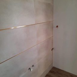 „Usługi Ogólnobudowlane KRIBUD” - Wykończona ściana z beżowych płytek, przedzielonych złotymi listwami, w łazience. Widoczne przyłącza wodne i fragment drewnopodobnej podłogi.