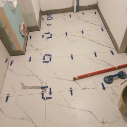 „Usługi Ogólnobudowlane KRIBUD” - Układanie białych płytek imitujących marmur na podłodze w łazience, z użyciem niebieskich klipsów poziomujących i poziomicy. Widoczny fragment ściany z płytą karton gips.