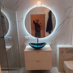 „Usługi Ogólnobudowlane KRIBUD” - Nowoczesna łazienka z marmurowymi płytkami, okrągłym lustrem z podświetleniem LED,  szklaną kabiną prysznicową i designerską umywalką.