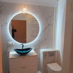 „Usługi Ogólnobudowlane KRIBUD” - Elegancka łazienka po remoncie: umywalka z czarną misą, podświetlane lustro w okrągłej ramie, marmurowe płytki z złotymi akcentami i podwieszana toaleta.