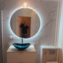 „Usługi Ogólnobudowlane KRIBUD” - Wykończona łazienka z umywalką w kształcie misy, okrągłym lustrem z podświetleniem LED i płytkami imitującymi marmur z złotymi akcentami.