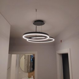 „Usługi Ogólnobudowlane KRIBUD” - Nowoczesna lampa sufitowa z okręgami LED w szarym wnętrzu. Widoczny fragment łazienki z lustrem i umywalką. Minimalistyczny design i ciepłe oświetlenie.