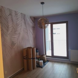 „Usługi Ogólnobudowlane KRIBUD” - Pokój po malowaniu z wzorem liści na ścianie, drewniana podłoga, lampa wisząca i balkon. Widoczne materiały malarskie przy ścianie.