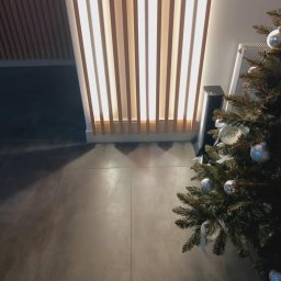 „Usługi Ogólnobudowlane KRIBUD” - Nowoczesna aranżacja wnętrza z podświetlaną ścianą z drewnianych paneli, szare płytki podłogowe i fragment choinki z srebrnymi bombkami w rogu.