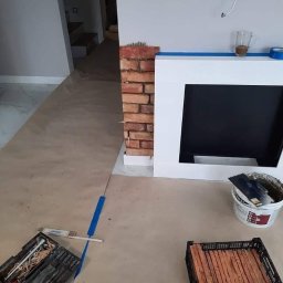 „Usługi Ogólnobudowlane KRIBUD” - Montaż cegły dekoracyjnej przy kominku w Choczewie. Widoczne narzędzia glazurnicze, płytki i zabezpieczone podłoże.