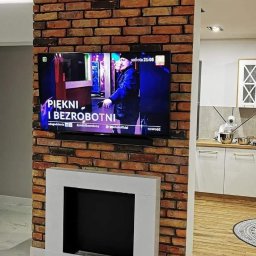 „Usługi Ogólnobudowlane KRIBUD” - Ściana z cegły z telewizorem i kominkiem w nowoczesnym salonie z błyszczącą podłogą. Widok na kuchnię w tle. Oświetlenie LED podkreśla strukturę cegły.