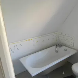 Biała wanna w łazience na poddaszu, ściany przygotowane do położenia płytek, widoczne ślady po kleju i zaznaczony kształt grzejnika.