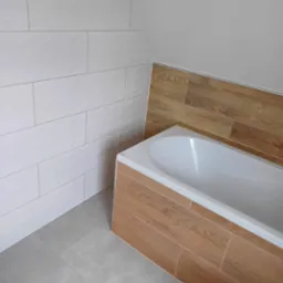 Wykończona łazienka na poddaszu: białe płytki ścienne, drewnopodobna obudowa wanny, szare płytki podłogowe. Ukośny sufit.