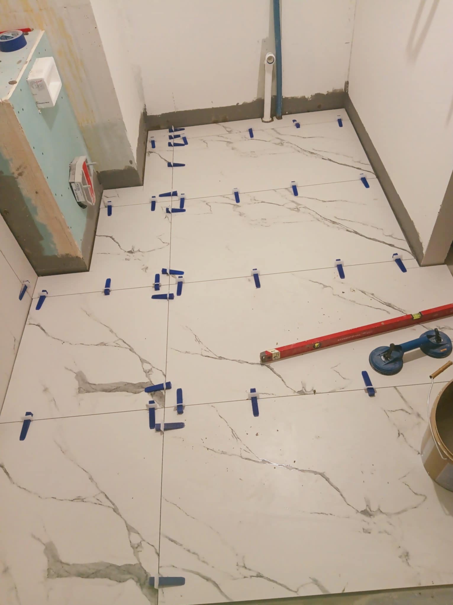 Układanie białych płytek imitujących marmur na podłodze w łazience, z użyciem niebieskich klipsów poziomujących i poziomicy. Widoczny fragment ściany z płytą karton gips.