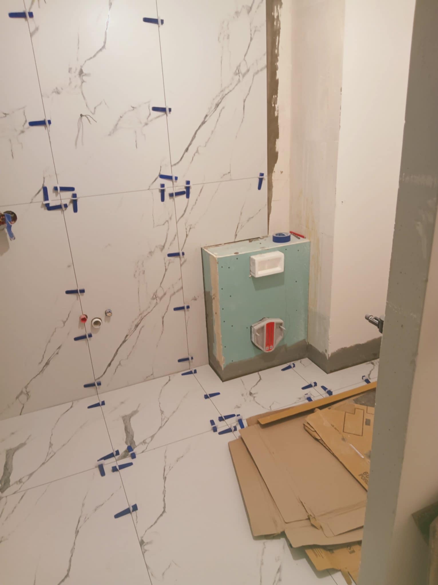 Układanie płytek marmuropodobnych w łazience, widoczne krzyżyki dystansowe i pionujące sznurki. Częściowo obudowany stelaż podtynkowy WC.