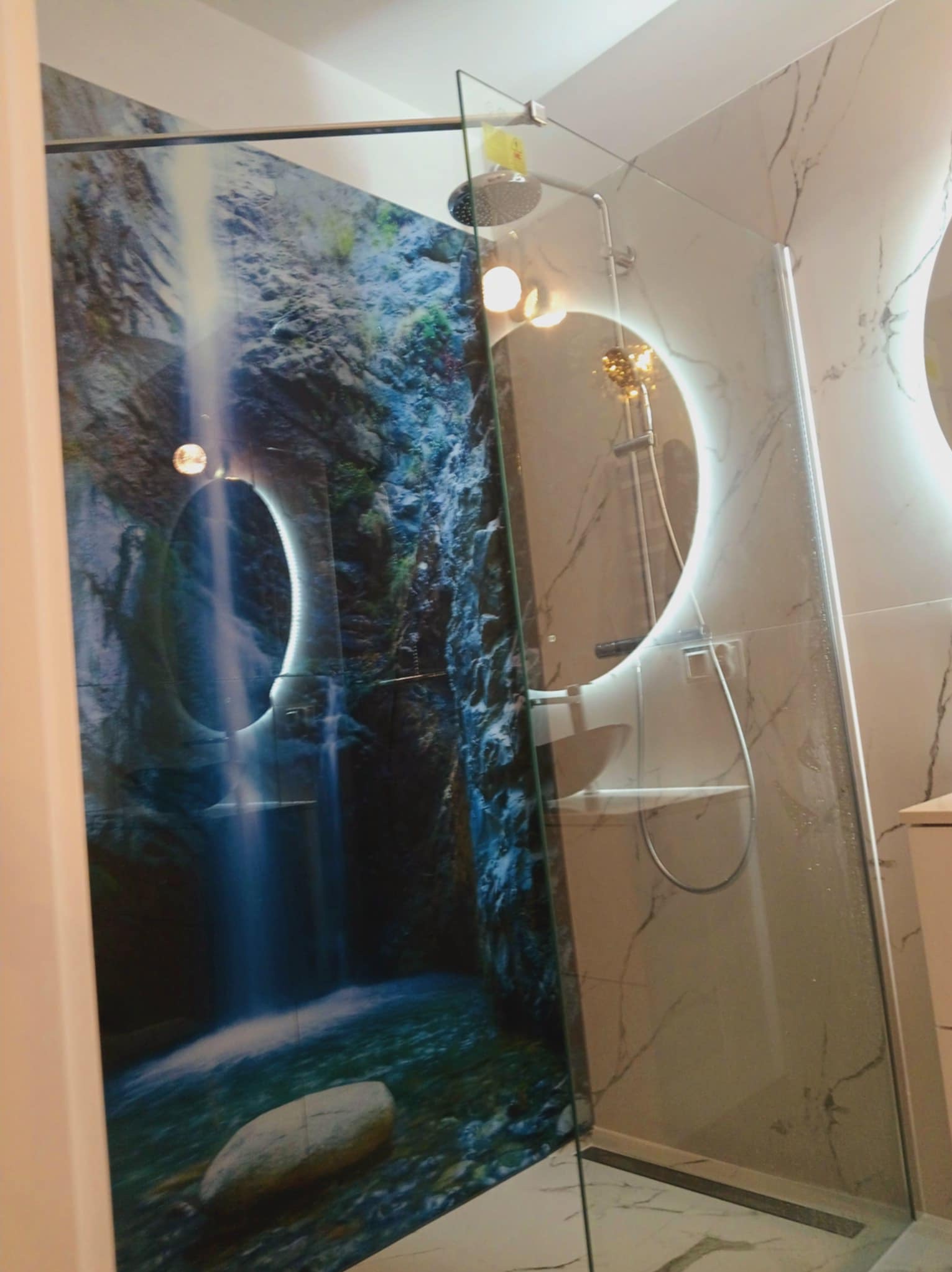 Nowoczesna kabina prysznicowa z grafiką wodospadu na szkle i marmurowymi płytkami, okrągłe lustro z podświetleniem LED, jasne wnętrze łazienki.