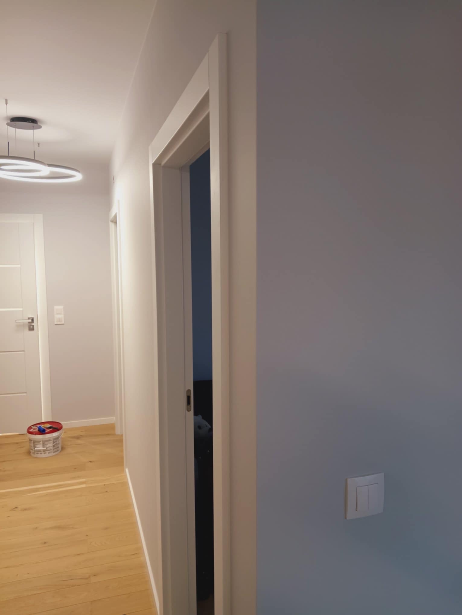 Jasne wnętrze po remoncie: pomalowane ściany w odcieniach szarości, drewniana podłoga, białe drzwi z ościeżnicami. Widoczny włącznik światła i lampa sufitowa.