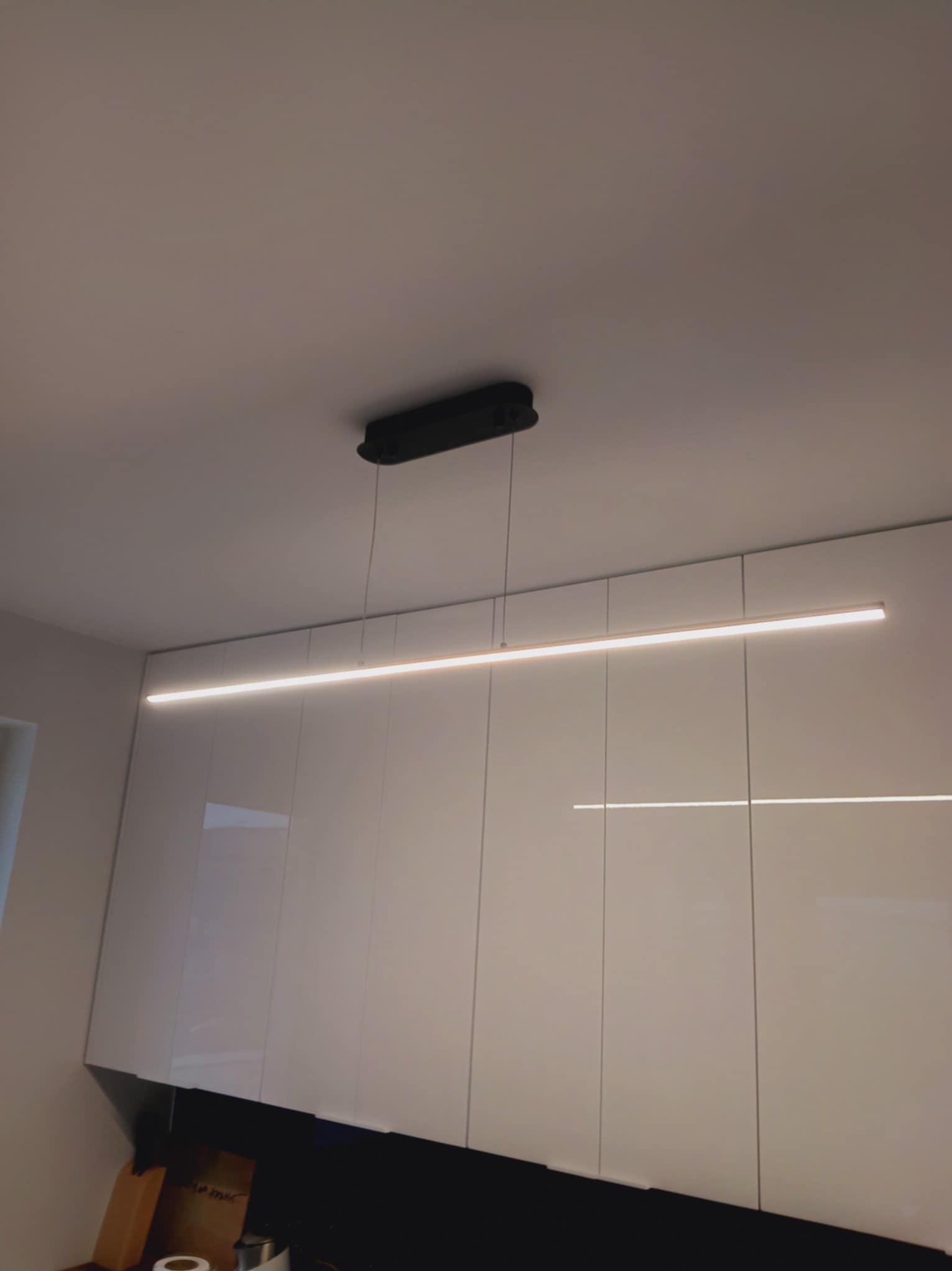 Minimalistyczna kuchnia z białymi, lakierowanymi szafkami podwieszanymi i nowoczesnym oświetleniem LED. Długa lampa sufitowa z czarną obudową.