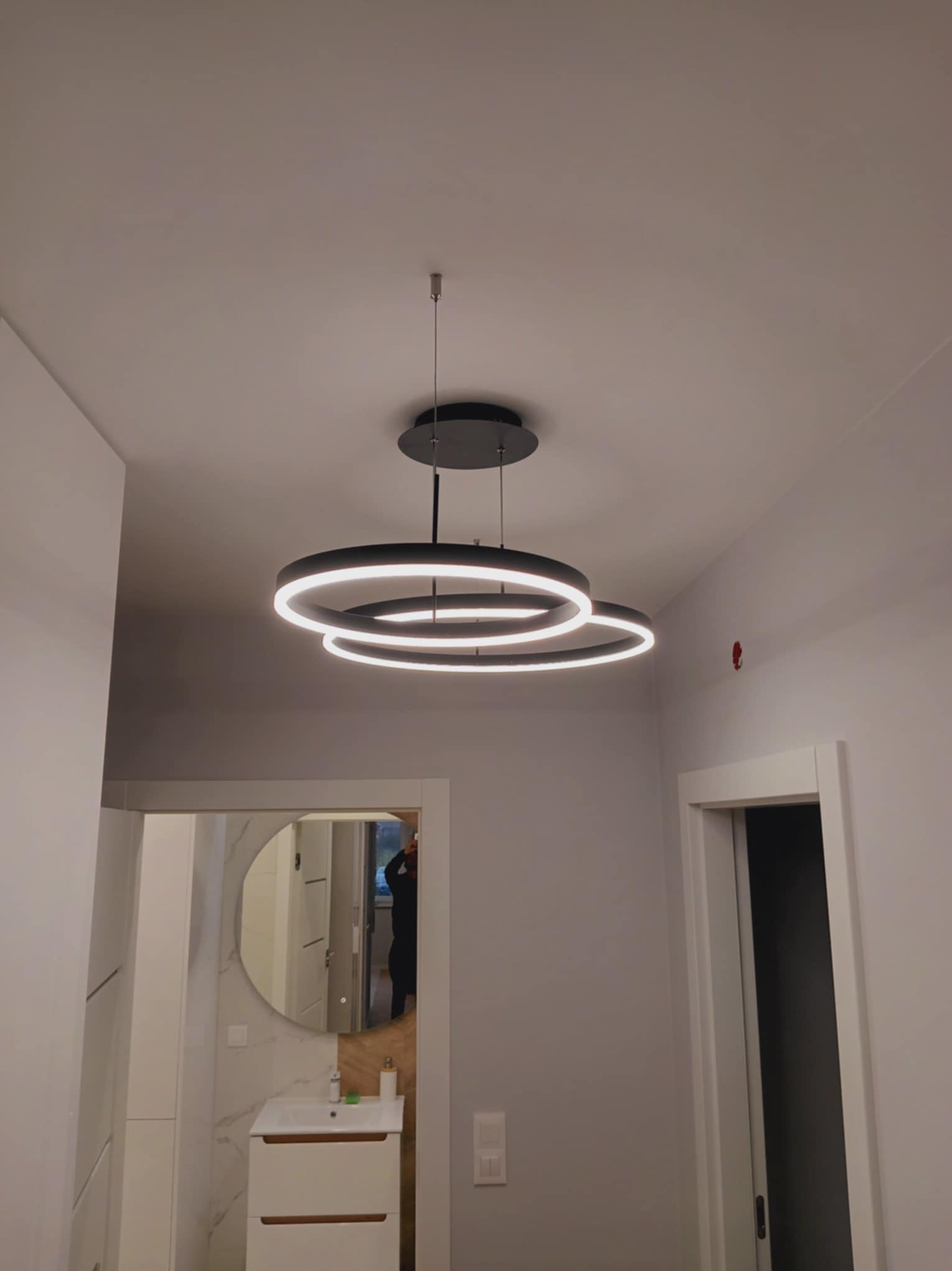 Nowoczesna lampa sufitowa z okręgami LED w szarym wnętrzu. Widoczny fragment łazienki z lustrem i umywalką. Minimalistyczny design i ciepłe oświetlenie.