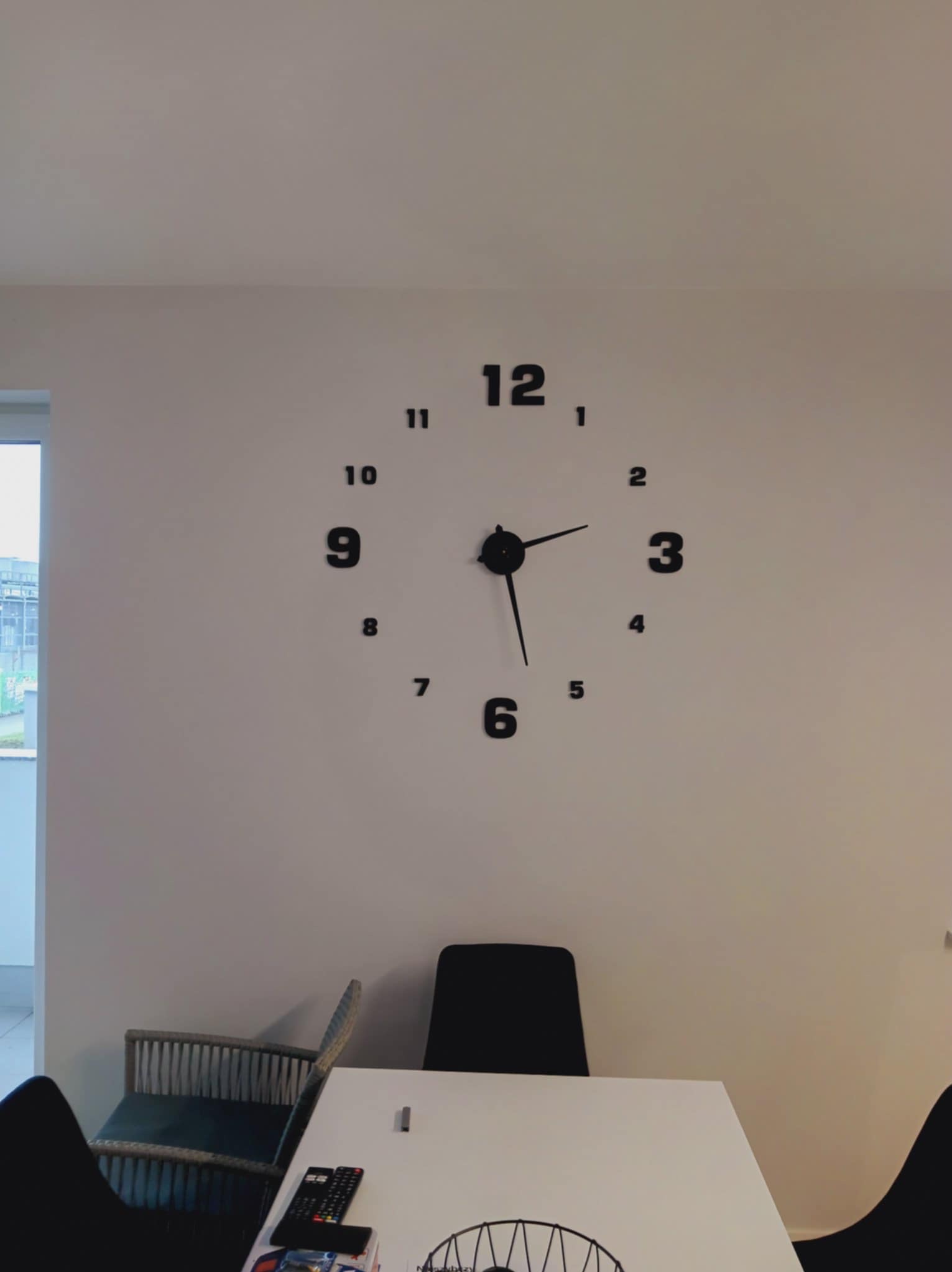 Minimalistyczny salon z dużym, samoprzylepnym zegarem na ścianie. Białe ściany, nowoczesne krzesła i stół tworzą spójną, jasną przestrzeń.