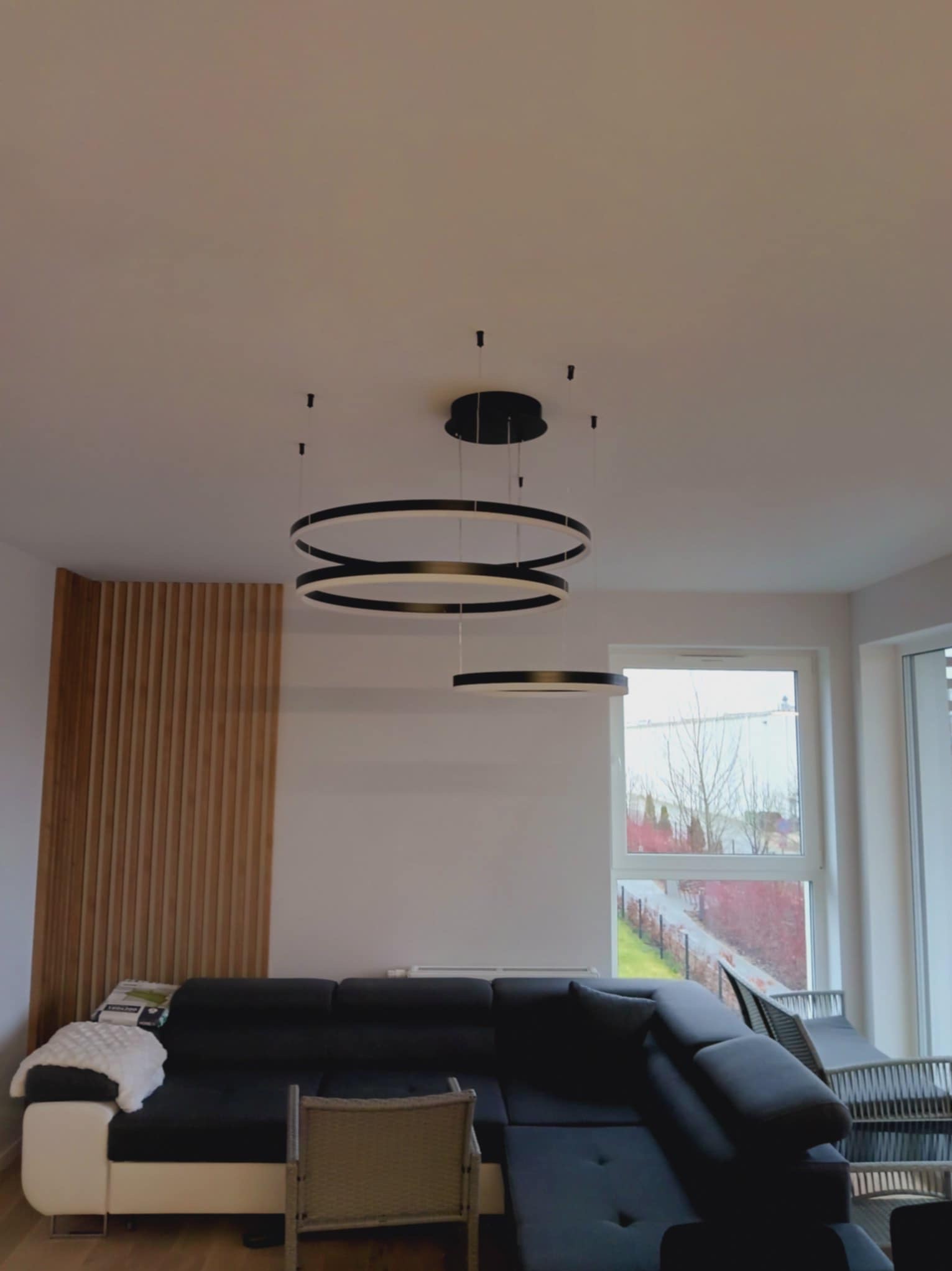 Nowoczesny salon z czarnym narożnikiem i dekoracyjną ścianą z drewnianych listew. Okrągła, wisząca lampa LED dodaje elegancji wnętrzu z widokiem na zewnątrz.