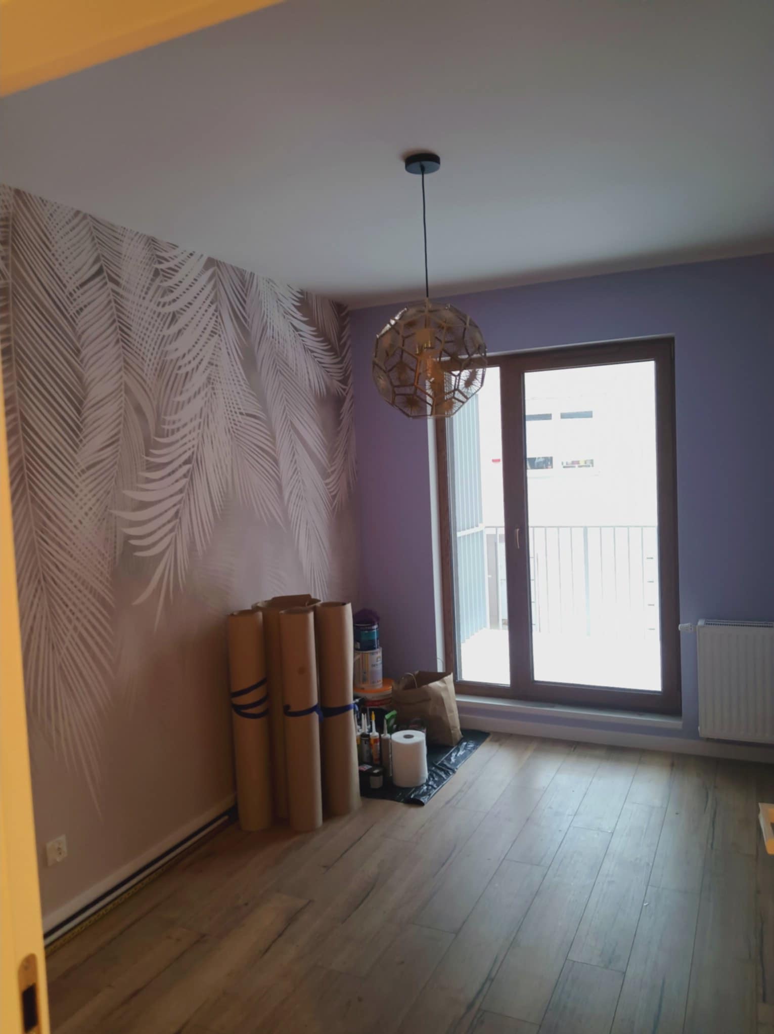 Pokój po malowaniu z wzorem liści na ścianie, drewniana podłoga, lampa wisząca i balkon. Widoczne materiały malarskie przy ścianie.